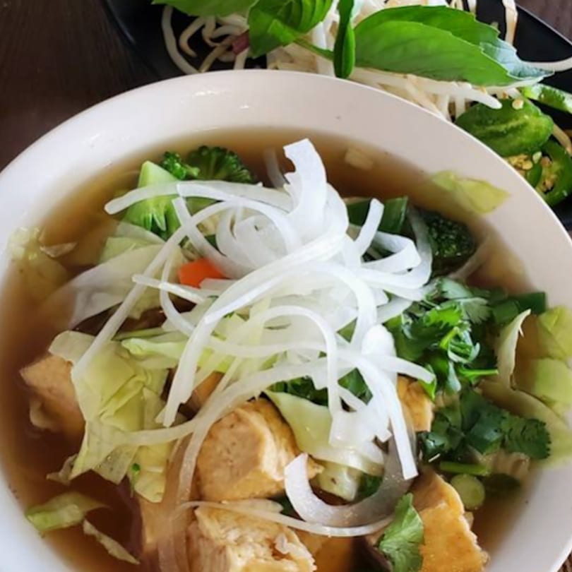 A bowl of Vietnamese pho with tofu, cabbage, broccoli, and sliced onions, with a side of basil and jalapeños.